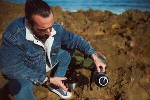Bearded male tourist traveler pouring water from a touristic thermos into a cup, sitting on the rocky cliff in by ocean photo