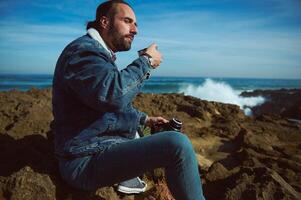 Relaxed male tourist refreshing himself with hot herbal drink, sitting on a rocky beach by ocean on a sunny autumn day photo