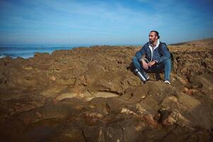 Young bearded man traveler, sitting alone on the rocky cliff, looking into the distance, enjoying the nature photo