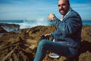 Happy male tourist smiling looking at camera, resting by sea, drinking refreshing drink against splashing ocean waves photo