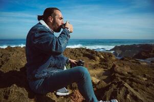 Contented traveler tourist sitting on the cliff, resting by sea, drinking refreshing drink against splashing ocean waves photo