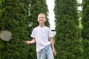 A happy boy holds a badminton racket in his hands, playing sports outside in summer photo