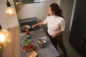 View from above of young adult woman in the minimalist home kitchen cooking dinner, putting a frying pan on the electric stove on induction oven photo