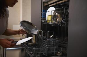 Close-up woman, housewife loading dirty dish inside a dishwasher, doing housework at home kitchen interior with minimalist design. Household. Chores. Housekeeping. Domestic life and routine photo