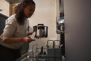 Multi ethnic woman, housewife unloading a clean stainless saucepan from a dishwasher. Modern kitchen appliances. Housework. Household. Chores. Housekeeping. Domestic life and routine photo