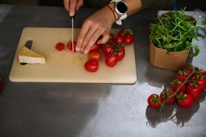 Overhead view of woman chopping tomato cherry on cutting board. A slice of delicious cheese, a bunch of organic tomatoes and fresh arugula leaves on the kitchen countertop. Italian Mediterranean food photo