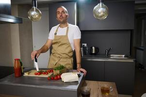 retrato de un caucásico medio Envejecido hombre, un cocinero en beige delantal, en pie a cocina mostrador con Fresco ingredientes, participación un cocina cuchillo y con confianza a cámara, Cocinando sano ensalada para cena foto