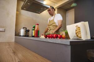 A bunch of red ripe organic tomato cherry and a packet with Italian pasta, against blurred male chef standing at induction stove, adjusting program while cooking dinner in modern home kitchen interior photo