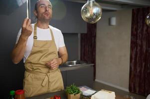 joven masculino cocinero saboreo ingredientes, besos dedos y haciendo delicioso dedo gesto mientras Cocinando cena en el hogar cocina foto