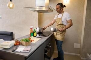 Handsome man in beige chef's apron cooking Italian pasta in the home kitchen, putting a saucepan on an electric stove to boil water for pasta, standing at kitchen table with fresh ingredients photo