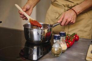 de cerca manos de un masculino cocinero participación un de madera espátula, emocionante tomate jugo con condimentos, Cocinando passata según a tradicional receta en el hogar cocina. italiano culinario. Mediterráneo comida foto