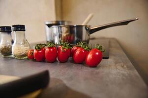 Still life with a branch of fresh ripe organic tomato cherry against blurred background of steel saucepan and bottles of condiments on the kitchen counter. Food background photo