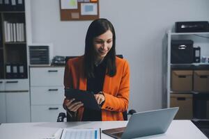 sonriente joven asiático mujer de negocios trabajando en un ordenador portátil computadora a su escritorio en un brillante moderno hogar oficina, haciendo calculador gastos financiero reporte Finanzas haciendo notas en papel grafico datos documento. foto