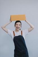 Smiling Asian woman in casual clothes holding a cardboard box mockup while standing against an isolated white background. shipping business concept photo