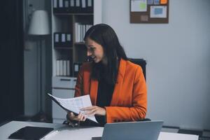 Smiling young Asian businesswoman working on a laptop computer at her desk in a bright modern home office, doing calculating expense financial report finance making notes on paper graph data document. photo