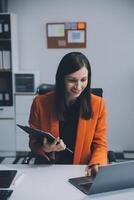 Smiling young Asian businesswoman working on a laptop computer at her desk in a bright modern home office, doing calculating expense financial report finance making notes on paper graph data document. photo