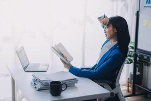 asiático mujer sensación migraña cabeza tensión.con exceso de trabajo mujer de negocios financiero mientras trabajando en ordenador portátil y tableta a moderno oficina. foto