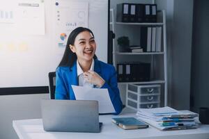 una alegre mujer de negocios que trabaja en una laptop en la oficina, una feliz y hermosa mujer de negocios asiática con traje formal trabaja en el lugar de trabajo. atractiva sonrisa de empleada de oficina. foto