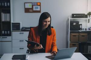 Smiling young Asian businesswoman working on a laptop computer at her desk in a bright modern home office, doing calculating expense financial report finance making notes on paper graph data document. photo