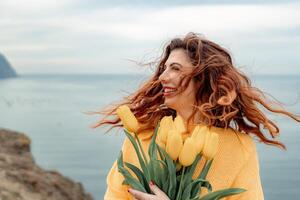 Portrait of a happy woman with hair flying in the wind against the backdrop of mountains and sea. Holding a bouquet of yellow tulips in her hands, wearing a yellow sweater photo