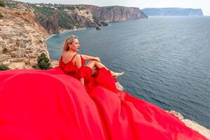 un mujer en un rojo seda vestir se sienta por el Oceano con montañas en el fondo, su vestir balanceo en el viento. foto