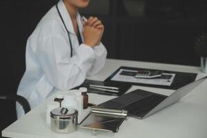 Serious female doctor using laptop and writing notes in medical journal sitting at desk. Young woman professional medic physician wearing white coat and stethoscope working on computer at workplace. photo