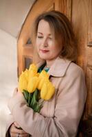 A woman is holding a bouquet of yellow tulips. She is wearing a tan coat and a green shirt. photo