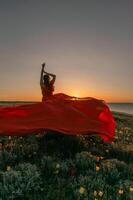 A woman in a red dress is standing in a field with the sun setting behind her. She is reaching up with her arms outstretched, as if she is trying to catch the sun. The scene is serene and peaceful. photo