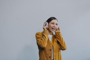 The beautiful emotional European brown-haired woman with healthy clean skin, charming smile, dressed in casual light clothes, listens and enjoys music on headphones with tablet on a white background. photo