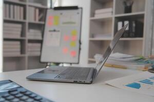 Close up view of simple workspace with laptop, notebooks, coffee cup and tree pot on white table with blurred office room background photo