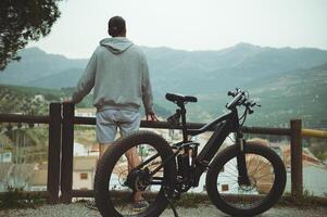 Rear view young active man with electric bicycle standing on top of hill and contemplating a beautiful view of a city in the mountains on foggy day. Healthy lifestyle. Sport. Inspiration. Achievements photo