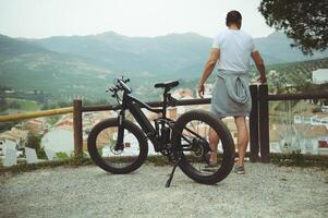 View from the back of a young man cyclist, standing near his electric bike, having a break after biking in mountains. People. Active healthy lifestyle. E-bike as sustainable, eco friendly transport photo