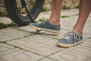 Cropped view male legs in denim sneakers near the electric bicycle wheel. photo