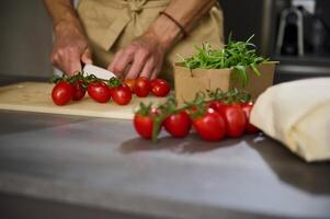 Close-up view of a male chef's hands chopping tomato cherry on a cutting board. Ingredients on the kitchen table. Man cooking healthy salad in the minimalist home kitchen interior photo