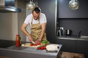 hermoso joven hombre en beige del chef delantal, Cocinando sano vegetariano ensalada en el moderno minimalista hogar cocina, en pie a cocina isla y el cortar Fresco maduro Cereza Tomates. foto