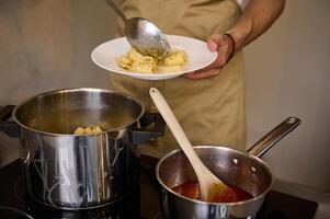 Close-up male chef plating up freshly cooked pasta, putting boiled penne on a white plate, standing at induction electric stove with saucepan full of tomato pasta. Italian culinary. Food background photo