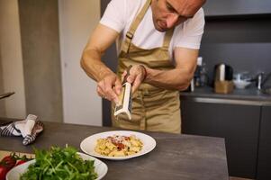 Close-up chef cooking in stylish modern minimalist home kitchen interior, grating parmesan cheese over a plate with freshly boiled pasta with tomato sauce. Ingredients on the kitchen countertop photo