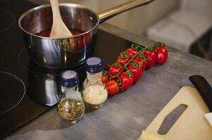 Two glass bottles of condiments, a branch of tomato cherry and steel pan with tomato sauce boiling and steaming on an electric stove in the home kitchen. Still life photo