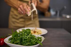 Closeup male chef cooking dinner at home, grating parmesan cheese on a pasta with tomato sauce. A bowl with fresh clean arugula leaves on the foreground photo