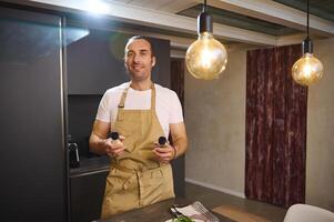 Caucasian male chef in beige kitchen apron, holding bottles of condiments for seasoning dish, standing at kitchen counter with fresh ingredients, looking at camera, preparing dinner at home kitchen photo