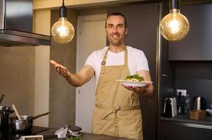 Smiling European man in beige chef's apron, holding a plate with Italian spaghetti pasta and showing it at camera. standing modern home kitchen interior. People and culinary photo