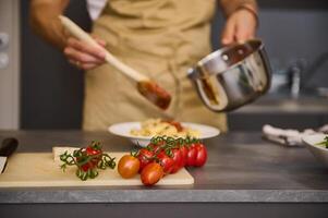 A branch of fresh ripe organic tomato cherry on the cutting board against the backdrop of a blurred male chef cooking Italian pasta at home kitchen. Traditional Italian cuisine. Food background photo