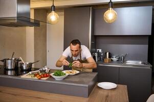 Male chef adding fresh green arugula leaves to Italian pasta with tomato sauce, garnishing the dish. A man preparing dinner in home kitchen. Selective focus on hands holding greens for seasoning food photo