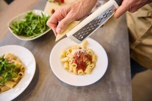 Directly above chef hands using grater, grating cheese on freshly cooked pasta with tomato sauce, seasoning and plating up the meal before serving it photo