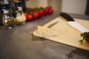 Selective focus on a slice of delicious savory parmesan cheese on cutting board, against blurred background of small bottles with condiments, a branch of cherry tomatoes on a gray kitchen countertop photo