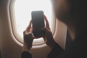 Blonde female tourist checking incoming notification on smartphone sitting on seat of airplane with netbook.Young businesswoman share media from telephone on laptop computer during plane flight photo