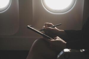Blonde female tourist checking incoming notification on smartphone sitting on seat of airplane with netbook.Young businesswoman share media from telephone on laptop computer during plane flight photo