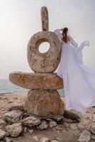 un mujer soportes en un Roca escultura hecho de grande piedras ella es vestido en un blanco largo vestido, en contra el fondo de el mar y cielo. el vestir desarrolla en el viento. foto