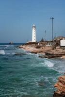 A lighthouse is on a rocky shoreline next to the ocean. The water is choppy and the sky is clear. photo