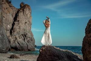 Woman white dress sea stones rocks.Middle-aged woman looks good with blond hair, boho style in a white long dress on beach jewelry around her neck and arms. photo